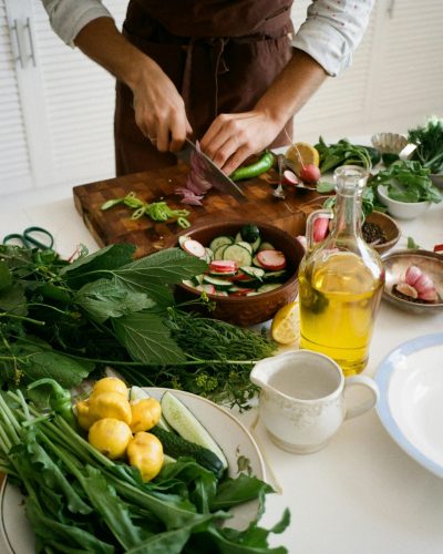 Adult preparing salad with fresh vegetables, knife, and olive oil in kitchen setting.